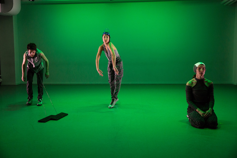 The space is cast in green as three dancers mingle in the space. One has two strings attached to his neck that are connected to computer keyboards